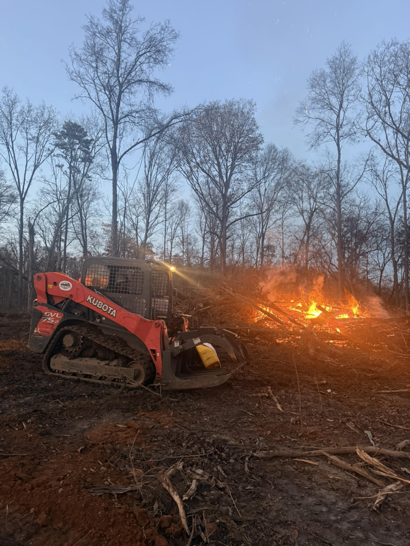 Kubota skid steer with grapple piling and burning brush during land clearing at dusk